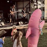 Children enjoy inflatable shark toys at a lively outdoor event as a band plays on stage, creating a festive atmosphere.