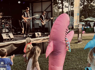 Children enjoy inflatable shark toys at a lively outdoor event as a band plays on stage, creating a festive atmosphere.