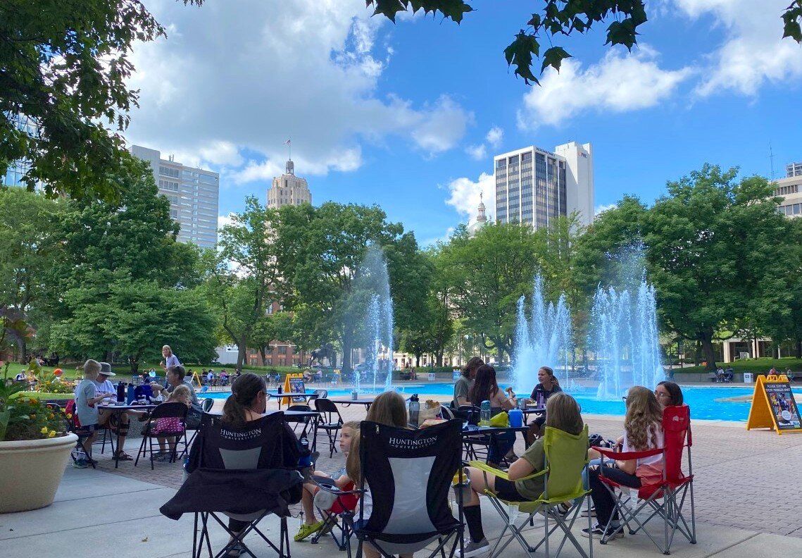 A vibrant park scene with people in chairs by a fountain, surrounded by trees and buildings under a sunny blue sky.