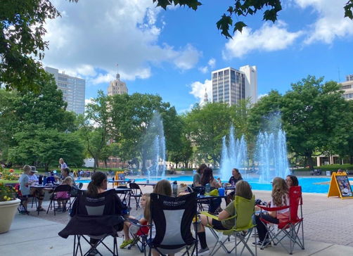 A vibrant park scene with people in chairs by a fountain, surrounded by trees and buildings under a sunny blue sky.