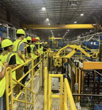 Group in safety vests and hard hats observes machinery on a platform, showcasing curiosity and engagement in a high-tech setting.