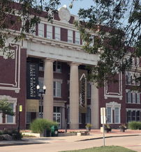 Classic brick building with tall columns, decorative details, surrounded by greenery and a blue sky, evoking tradition and stability.