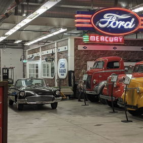 Vintage cars in a museum setting, featuring a classic black car, colorful trucks, and a neon Ford sign, evoke nostalgia.