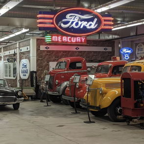 Vintage cars in a museum setting, featuring a classic black car, colorful trucks, and a neon Ford sign, evoke nostalgia.