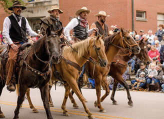 Cowboys in traditional Western attire ride horses in a lively parade, surrounded by an excited crowd celebrating community spirit.