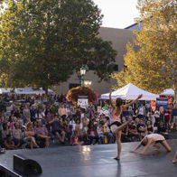 Dynamic dancers perform on stage at an outdoor festival, captivating an engaged audience amidst vibrant autumn foliage.