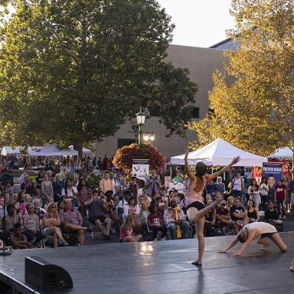 Dynamic dancers perform on stage at an outdoor festival, captivating an engaged audience amidst vibrant autumn foliage.