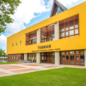 A vibrant yellow building named "Tubman," a welcoming art center surrounded by green grass and trees, evokes creativity and community.