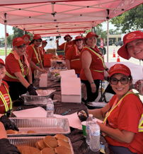 A cheerful group in red shirts and hats gathers under a pink tent, enjoying food and community spirit at an event.