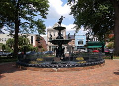Charming public square with an ornate fountain, surrounded by trees and flowers, evoking tranquility in a bustling town.