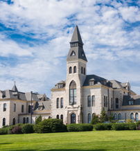 Historic building with a tall tower, mixed architecture, surrounded by lush greenery and a bright sky, evoking nostalgia and serenity.