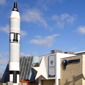 Modern building with rockets, sleek design, large windows, bright blue sky, evokes wonder and excitement for space exploration.