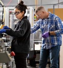 Two professionals in a lab: a woman focused on a machine and a man nearby, both in safety gear, reflecting teamwork and concentration.