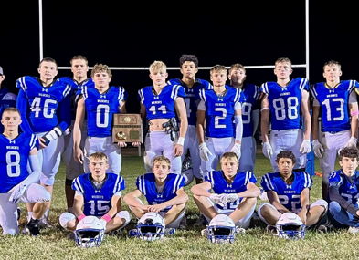 Young athletes in blue jerseys pose proudly on the field under stadium lights, celebrating unity and victory with a trophy.