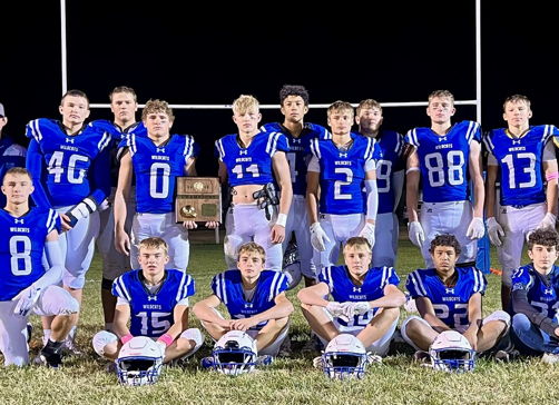 Young athletes in blue jerseys pose proudly on the field under stadium lights, celebrating unity and victory with a trophy.