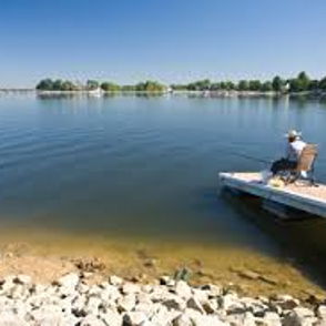 A person fishes peacefully from a wooden dock on a calm lake, surrounded by greenery under a clear blue sky.