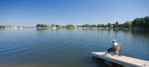 A person fishes peacefully from a wooden dock on a calm lake, surrounded by greenery under a clear blue sky.
