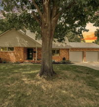 Charming brick house with a lush lawn, large tree, and a colorful sunset, evoking comfort and tranquility.