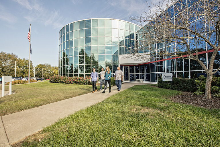 Three people walk on a path to a modern glass building, surrounded by greenery, reflecting a welcoming and professional atmosphere.