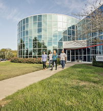 Three people walk on a path to a modern glass building, surrounded by greenery, reflecting a welcoming and professional atmosphere.