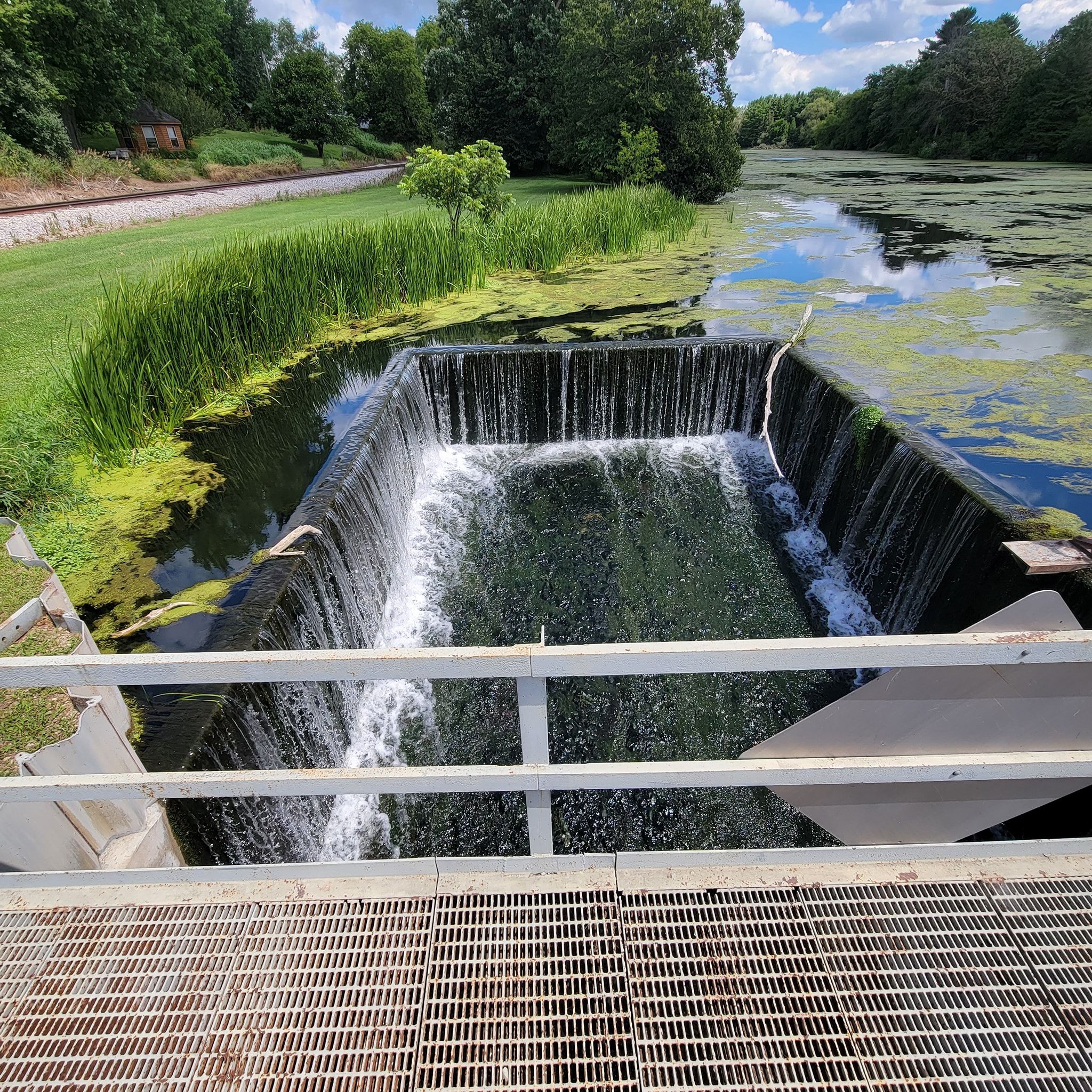 Tranquil waterway with a small waterfall, surrounded by lush greenery and reflections, evoking calm and serenity.