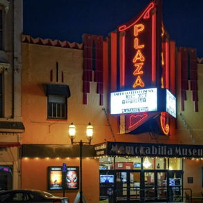 A vintage theater at night, illuminated with a red marquee "PLAZA," evoking a cozy, nostalgic community atmosphere.