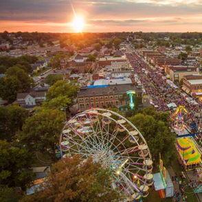 Aerial view of a vibrant festival at sunset, featuring a Ferris wheel, rides, and a joyful crowd, exuding celebration.