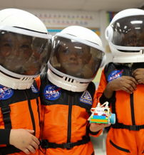 Three children in orange astronaut suits play with a toy rocket, expressing joy and curiosity in a vibrant, educational setting.
