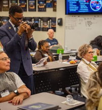 Classroom scene with engaged individuals; a man in a suit takes a photo, highlighting collaboration and a professional atmosphere.
