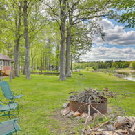 Cozy red house by tranquil water, surrounded by green trees and chairs around a fire pit, creating an inviting atmosphere.