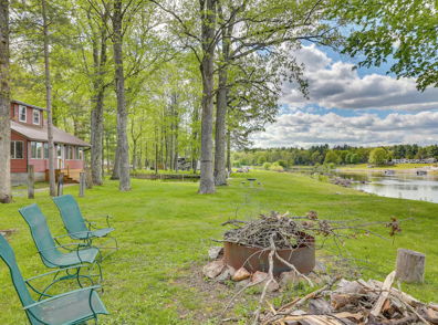 Cozy red house by tranquil water, surrounded by green trees and chairs around a fire pit, creating an inviting atmosphere.