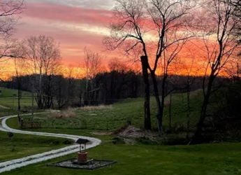 Sunset over a serene landscape with a winding path, lush green yard, and hills, evoking tranquility and reflection.