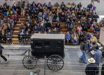 A lively indoor auction scene with a crowd around a showcased black horse-drawn carriage, evoking community and tradition.