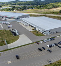 Aerial view of a large industrial facility with a flat roof, parking lot, surrounded by green hills and trees, conveying efficiency.