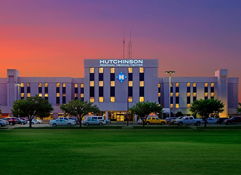 Hutchinson hospital at dawn/dusk, flanked by grass and trees, with a parking lot; warm sky evokes safety and hope.