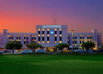 Hutchinson hospital at dawn/dusk, flanked by grass and trees, with a parking lot; warm sky evokes safety and hope.