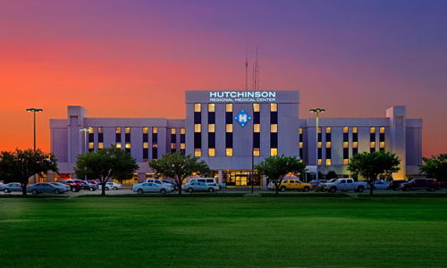 Hutchinson hospital at dawn/dusk, flanked by grass and trees, with a parking lot; warm sky evokes safety and hope.