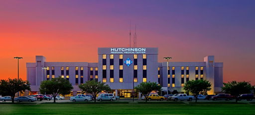 Hutchinson hospital at dawn/dusk, flanked by grass and trees, with a parking lot; warm sky evokes safety and hope.