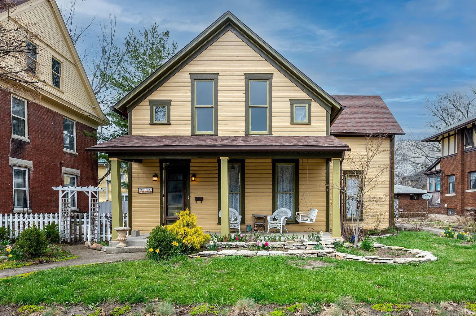 Charming yellow two-story house with dark trim, inviting porch, and lush yard, exuding warmth and tranquility.