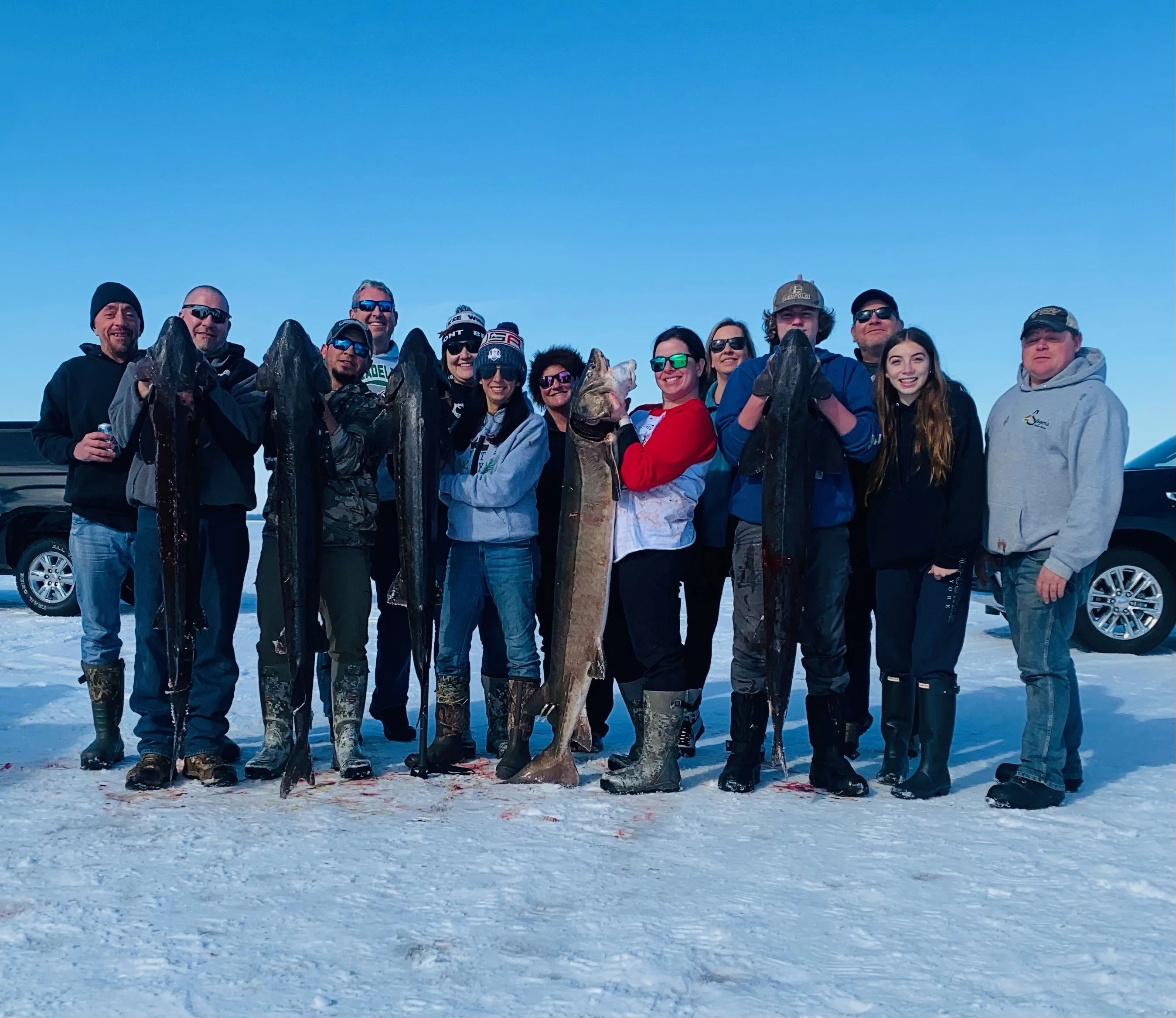Group joyfully displays large fish after a successful ice fishing trip in a snowy landscape against blue skies.