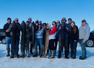 A group smiles and poses with large fish in a snowy landscape, celebrating camaraderie and success after a fishing trip.