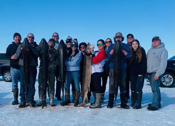 A group smiles and poses with large fish in a snowy landscape, celebrating camaraderie and success after a fishing trip.