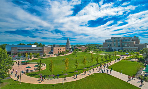 Sunny campus scene with grassy pathways, people strolling, trees, modern buildings, and a bright sky, exuding warmth and community.