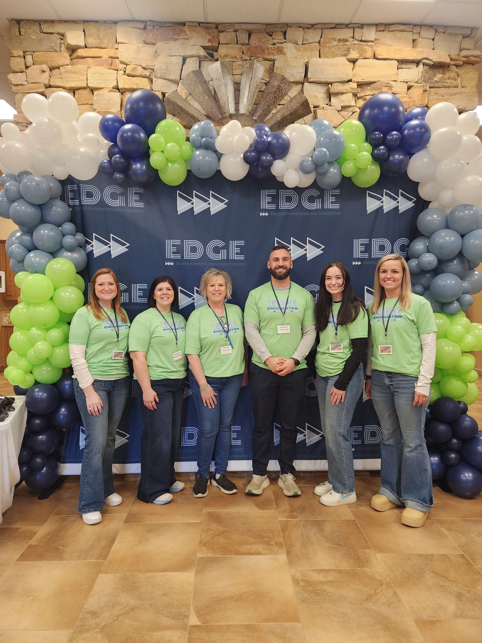 A group of six in matching green shirts stands in front of a colorful balloon backdrop, exuding celebration and teamwork.