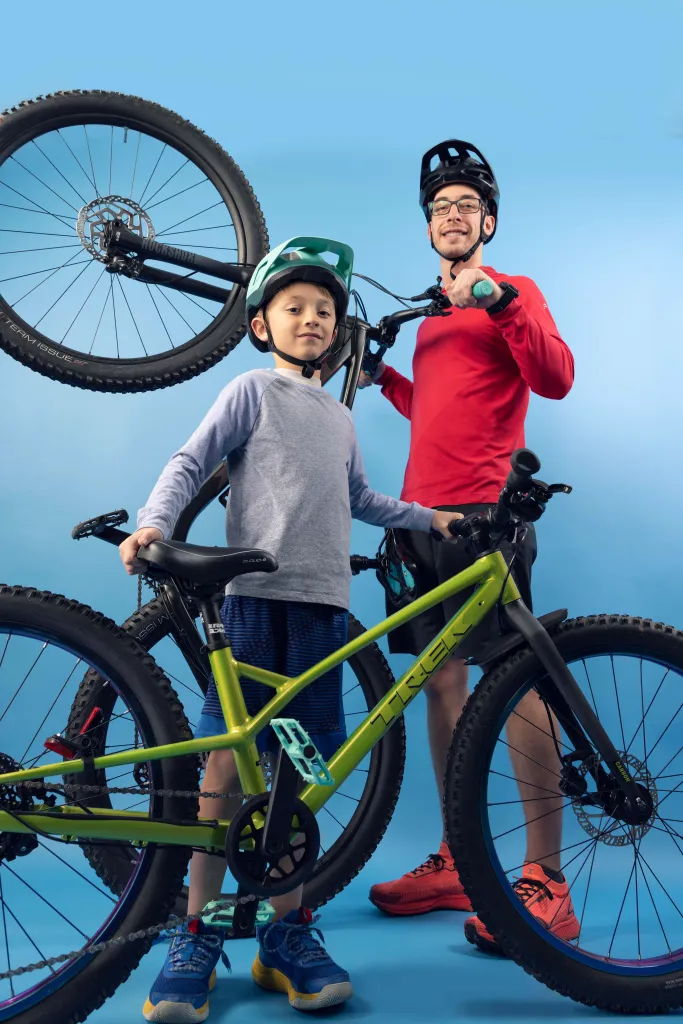 A man and boy in helmets pose with mountain bikes against a blue background, radiating joy and a love for adventure.