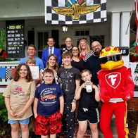 Group of people, including children and a racing mascot, joyfully pose in front of a race-themed decorated house.