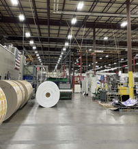 A spacious industrial facility with large material rolls and organized machinery, evoking productivity, simplicity, and innovation.