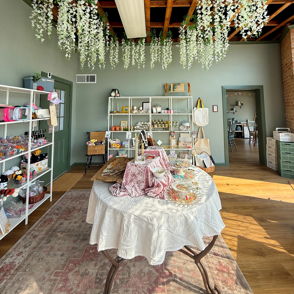 Charming shop interior with greenery, soft colors, rustic beams, and a welcoming table, evoking warmth and creativity.