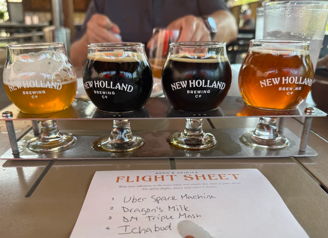 Four craft beer glasses in a wooden holder, showcasing lighter and darker brews from New Holland, with a tasting list.