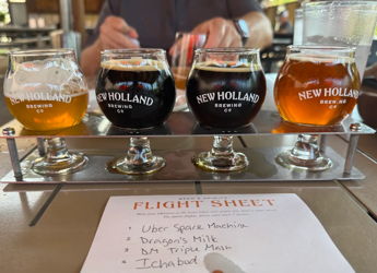 Four craft beer glasses in a wooden holder, showcasing lighter and darker brews from New Holland, with a tasting list.